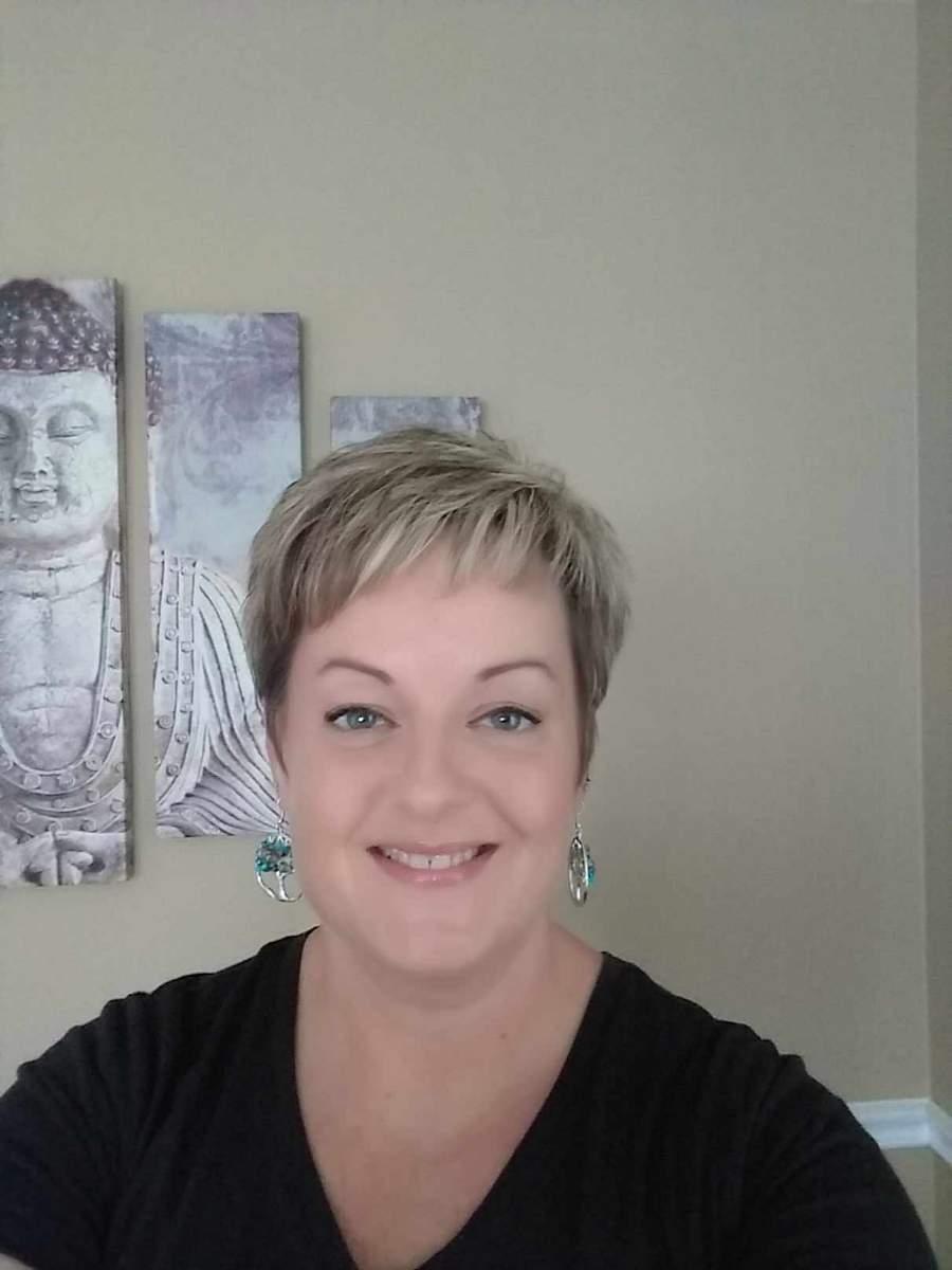 Julie is smiling standing in front of a picture of Buddha wearing earings made from stones in the image of the "Tree of Life".